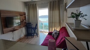A living area with a TV mounted on a wooden panel, a red sofa, a small table, and a potted plant. - Porto Real Resort - Daily Rates and Seasonal Rates (Mangaratiba)