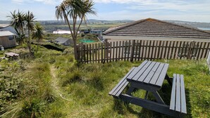 A wooden picnic table and bench in a grassy area with a view of a coastal village and hills in the background. - E42 Vista Marina, Riviere Towans (Hayle)