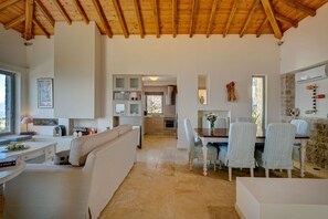 A spacious living area with a high wooden ceiling, a dining table with chairs, a kitchen in the background, and a cozy seating area with a sofa and a small table. - Villa Kalyvia Beta | Kalyvia, Skiathos | Private Infinity Pool & Sea Views (Skiathos)