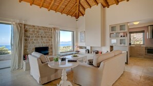Un'ampia zona giorno con parete in pietra, televisore, divano, tavolino da caffè e angolo cottura con mobili e finestra vista mare.