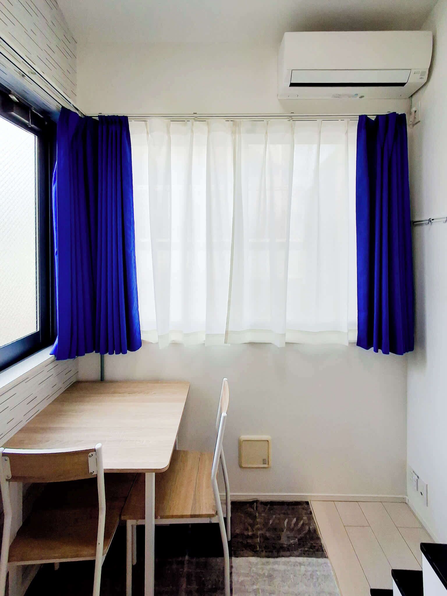 A small room with a wooden table and chairs, a window with white and blue curtains, and an air conditioning unit on the wall.