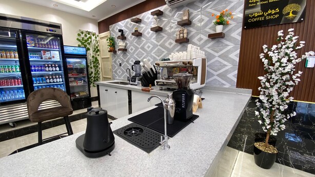 A coffee shop with a modern interior, a refrigerator stocked with beverages, and a coffee machine on the counter. - Rahet Al-Kharj Serviced Apartments (Al Kharj)