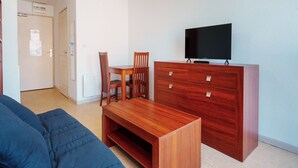 A modern living room with a wooden TV stand, a blue sofa, and a dining table with chairs. - Charming studio apartment for 2 people (Agde)