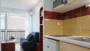 A compact kitchen with a microwave, a sink, and a refrigerator. There is a sofa and a small table in the living area. - Charming studio apartment for 2 people (Agde)
