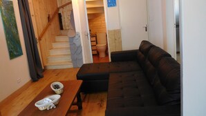 A living room with a black leather sofa, a wooden table with slippers, a staircase with a stone wall, and a view into a bathroom with a toilet and a blue wall.