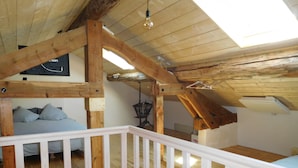 A loft-style bedroom with exposed wooden beams, a white railing, and a bed with grey bedding.