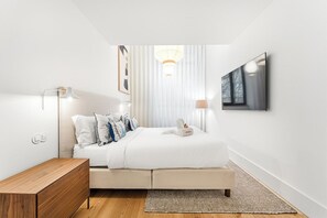 Een moderne slaapkamer met een bed, bedlampen, een houten nachtkastje, een aan de muur bevestigde televisie en een groot raam met dunne gordijnen.