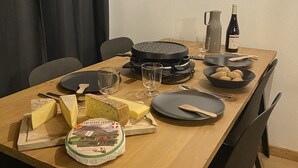 Een eettafel gedekt met borden, bestek, glazen, een fles wijn, een schaal aardappelen en een kaasplank.
