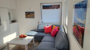 A modern living room with a grey sofa, a small white table with a bowl of apples, and a window with blinds. - Apartment 'Hanna' with Private Garden and Wi-Fi (Langeoog)