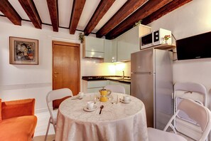 A dining area with a round table set for four, surrounded by white chairs. A microwave is mounted above the counter, and a refrigerator is visible to the right. - Emerald Apartment (Venezia)
