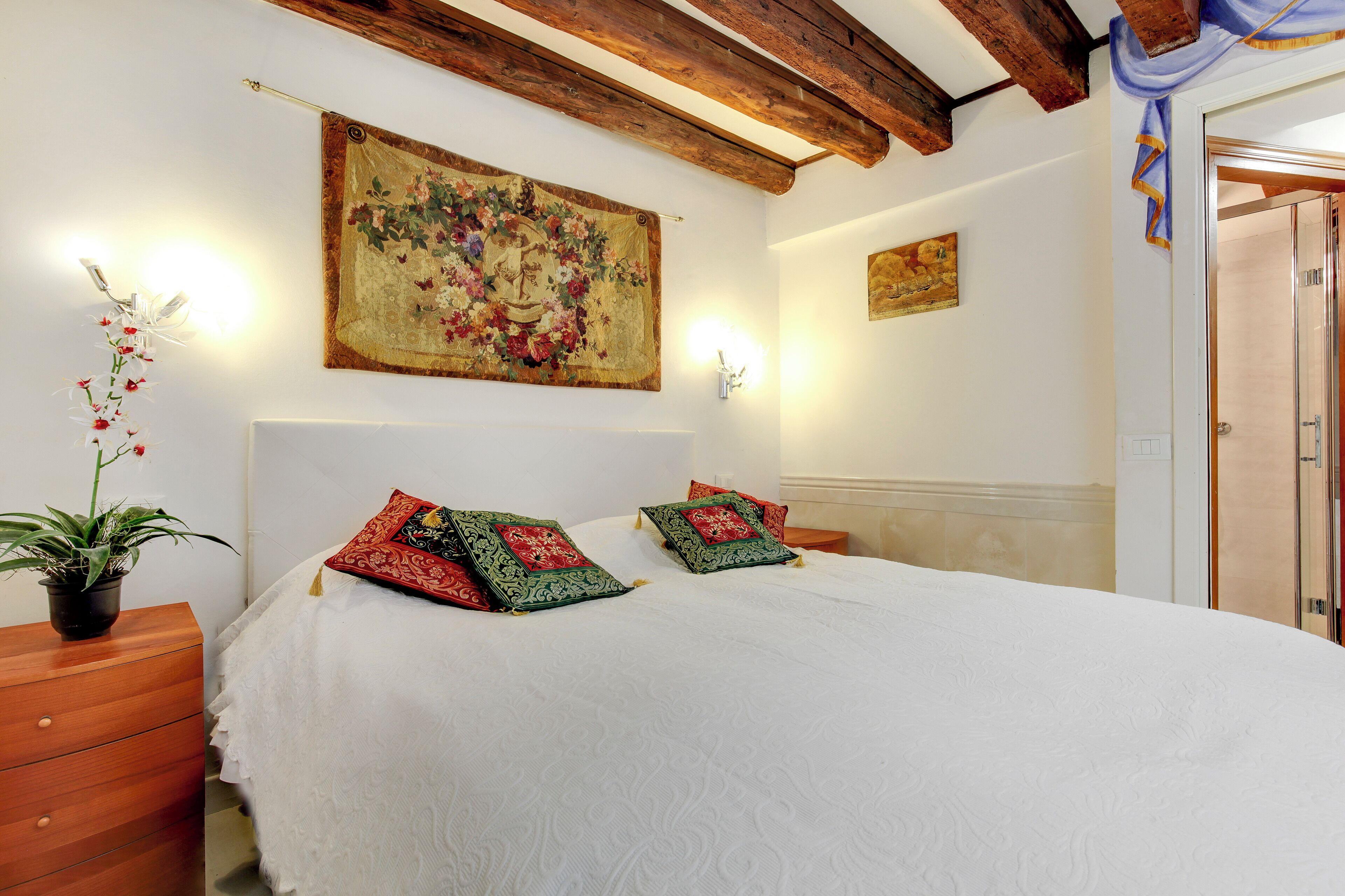 A bedroom with a large bed, two decorative pillows, a nightstand with a plant, and a painting on the wall.