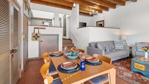 A dining area with a wooden table set for four, a staircase leading to an upper level, and a grey sofa in the living area. - Casa Golondrinas-A Serene Santa Fe Retreat (Santa Fe)