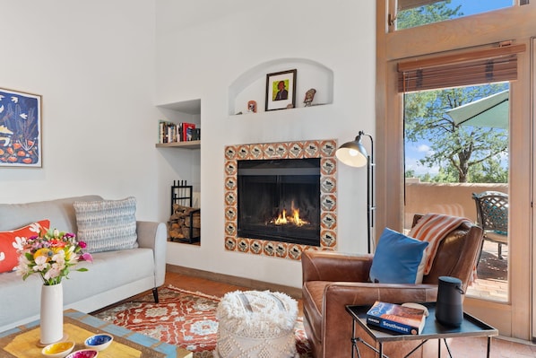 A living room with a fireplace, a sofa, a coffee table with a vase of flowers, and a view through a glass door.