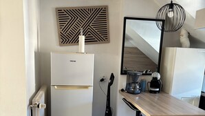 Et lille køkken med et hvidt køleskab, et træspisebord, en sort gulvlampe og en dekorativ vægdekoration.