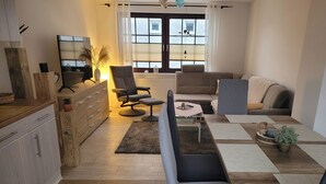 Una sala moderna con sofá, sillón y mesa de centro. Se observa un aparador y un espejo a la izquierda. En el fondo, una ventana con persianas.