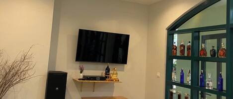 Una sala de estar moderna con un televisor de pantalla plana montado en la pared, una pequeña mesa con sillas, un altavoz y un mueble de vidrio con botellas.