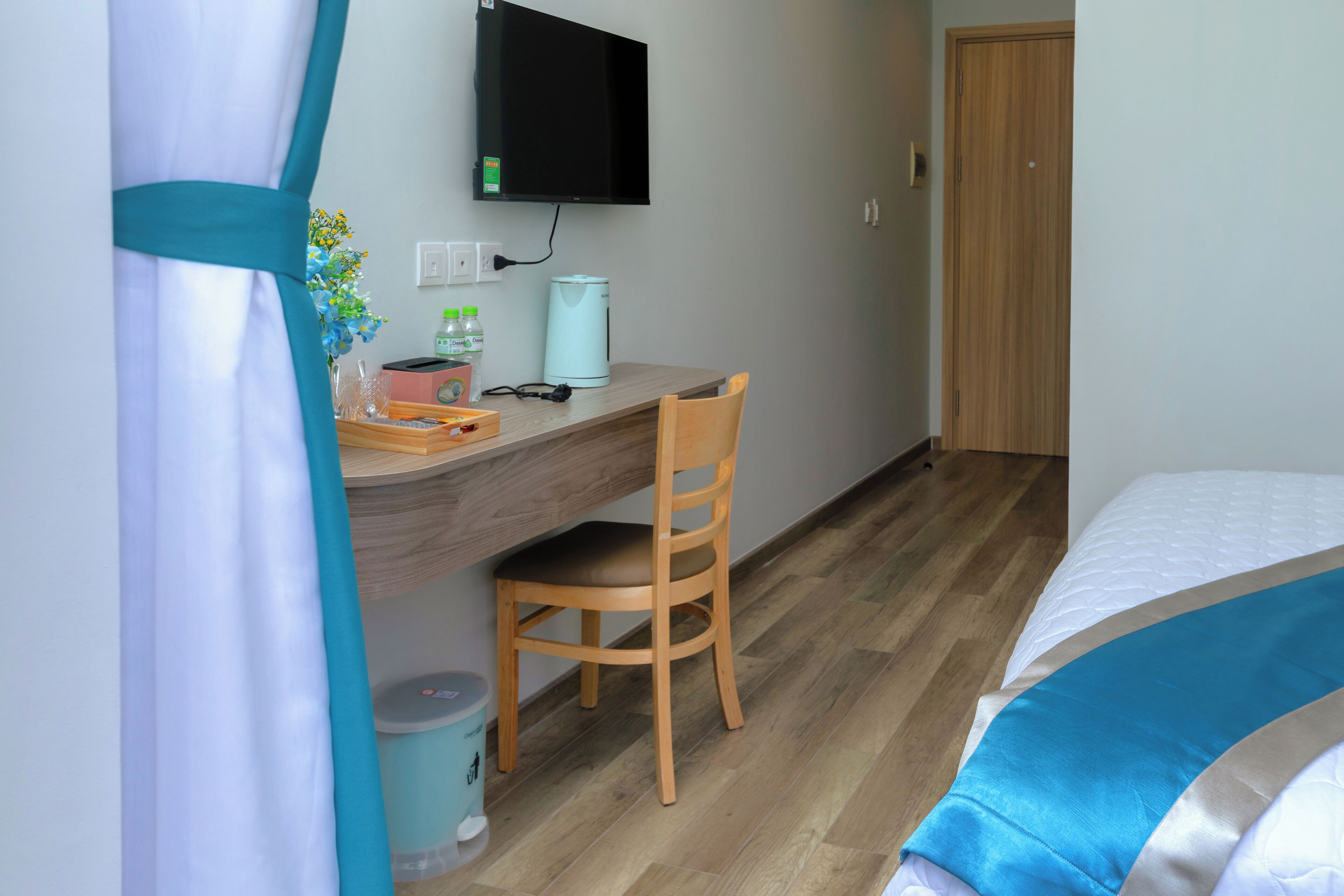 Une pièce avec un bureau en bois, une chaise, un lit recouvert d’une couverture bleue et blanche, un téléviseur fixé au mur et une porte en bois.