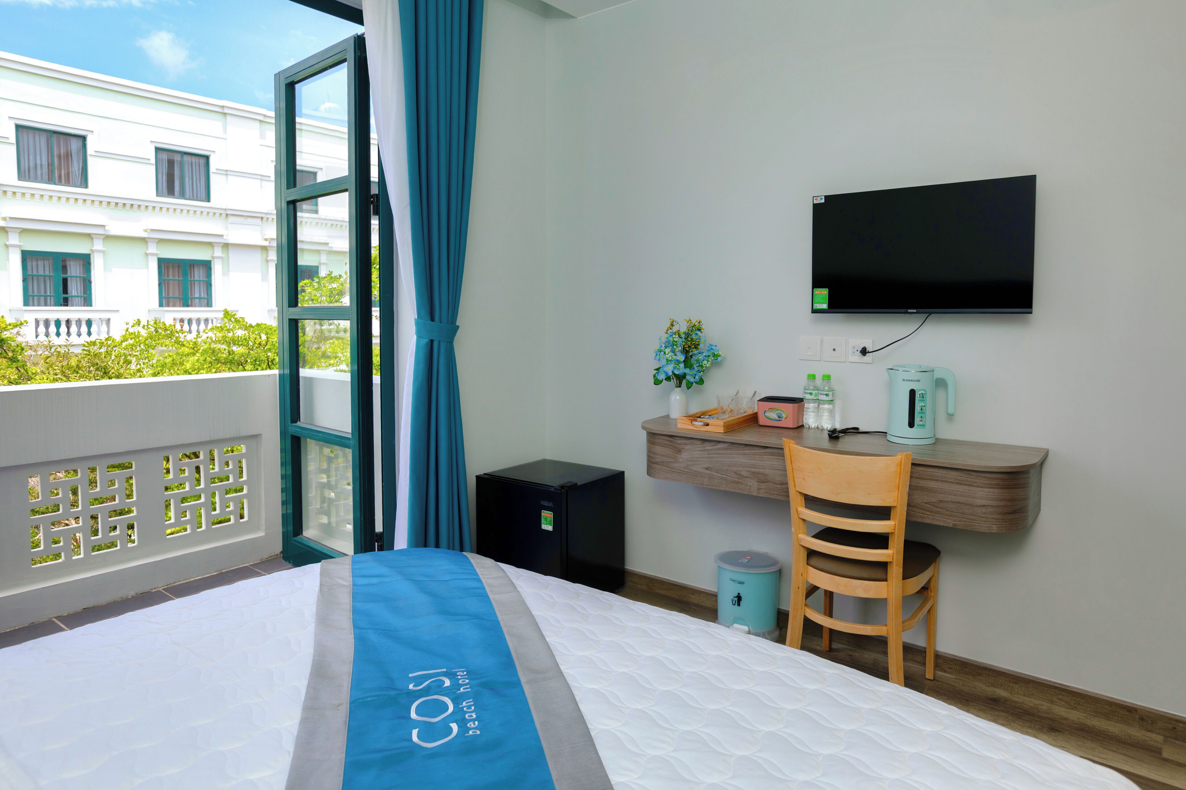 A hotel room with a bed, a desk with a chair, a TV, a small plant, and a trash bin.