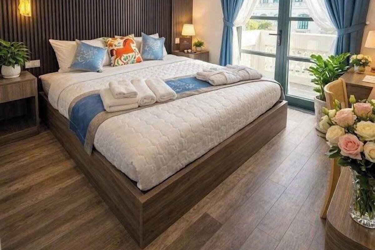 Une chambre d’hôtel moderne avec un grand lit, une tête de lit en bois, des lampes de chevet, une table de chevet, une fenêtre avec des rideaux et une vue sur la ville.