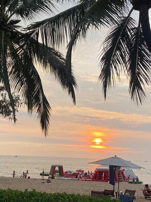 Une scène de plage avec des palmiers, un coucher de soleil, des structures gonflables pour jeux et des personnes se relaxant sur le sable.