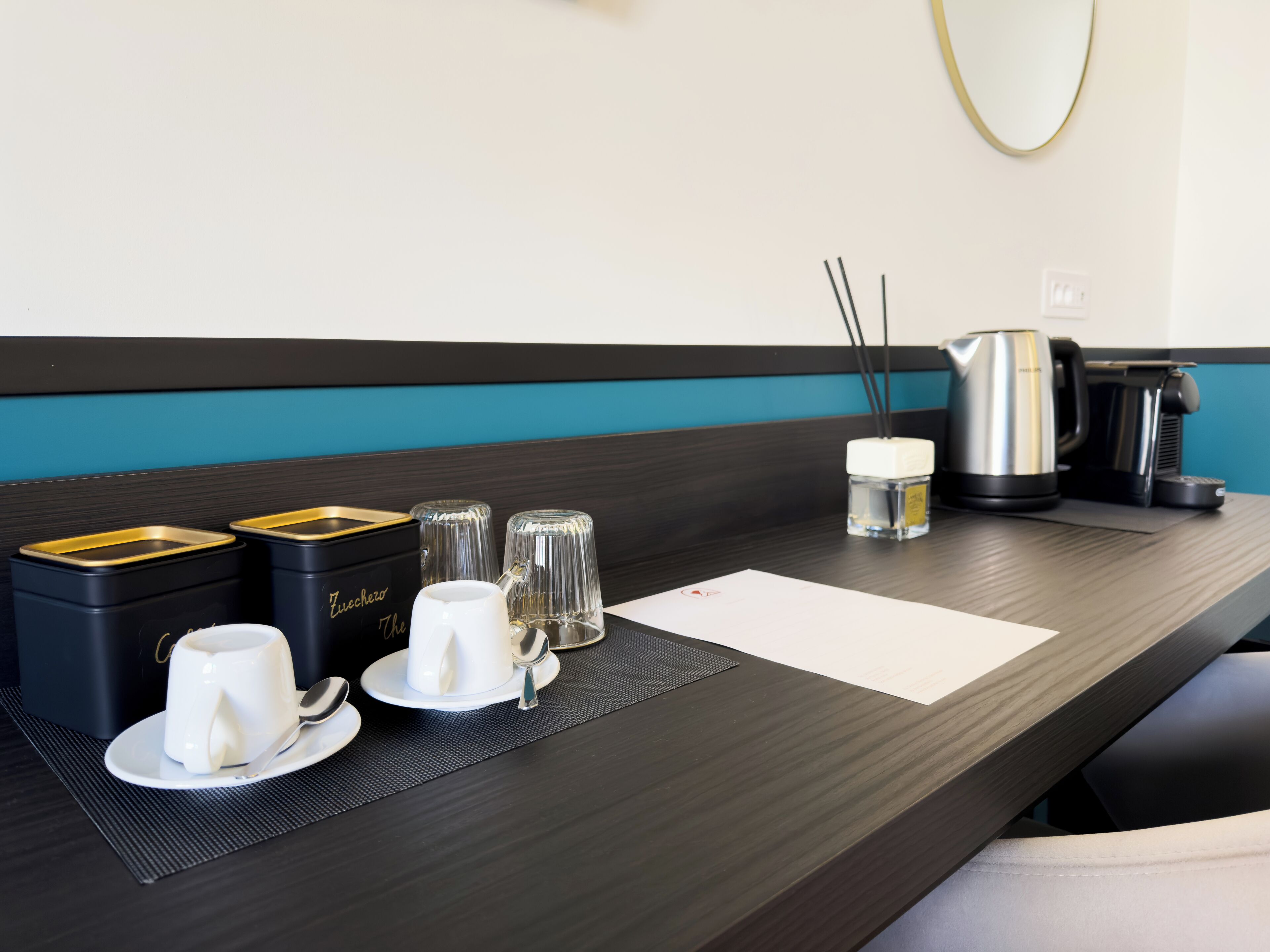 A hotel room with a dark wooden desk, a black and gold minibar, two white cups, two glass tumblers, a small bottle, a black and silver coffee maker, and a white paper on the desk.