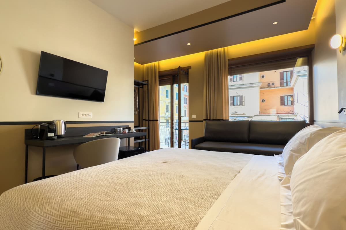 Habitación de hotel con cama, escritorio con cafetera, televisión y ventana con vista a edificios.