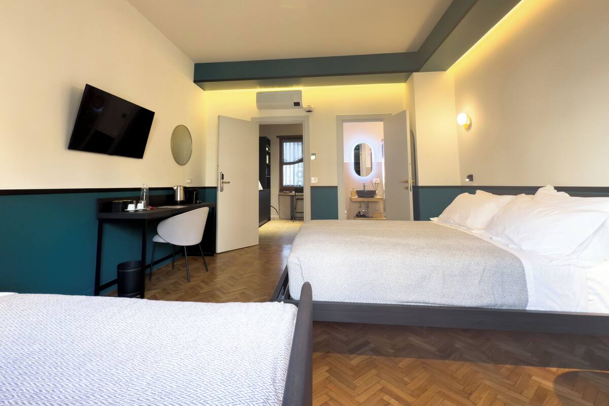 Habitación de hotel con dos camas, un escritorio, una silla, televisión, espejo y baño visible a través de una puerta abierta.
