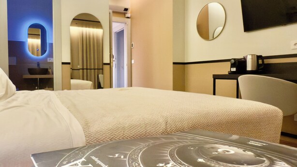 Ein Hotelzimmer mit einem großen Bett, einem Nachttischchen mit Kaffeemaschine, einem runden Spiegel an der Wand und einer Tür zu einem weiteren Raum.