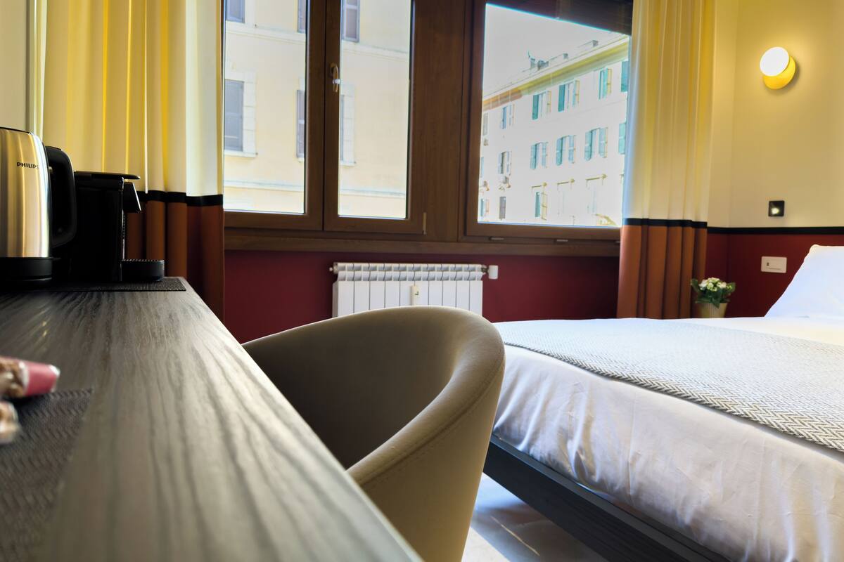 Habitación de hotel con cama, escritorio con cafetera, silla y ventana con vista a edificios.