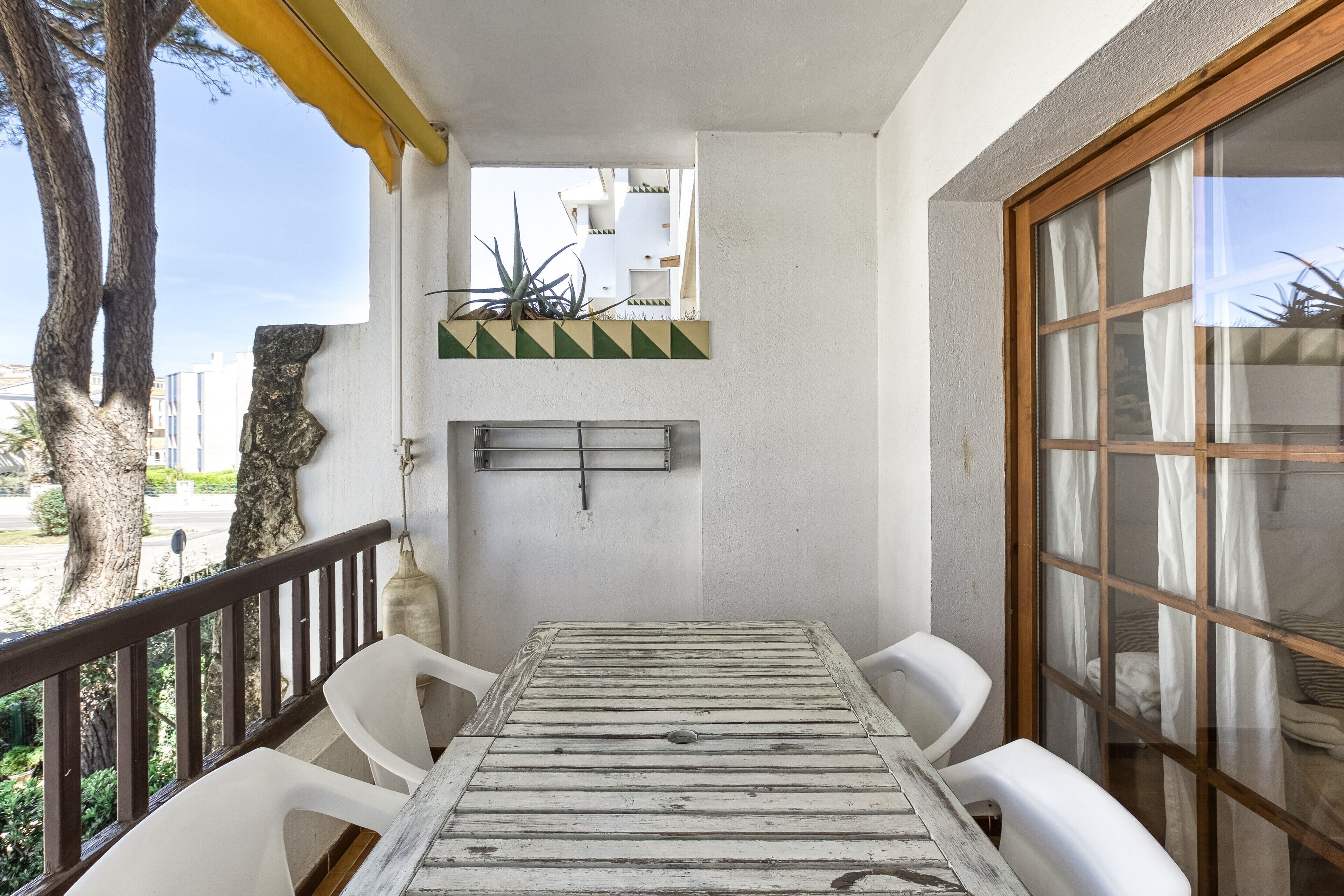 A balcony with a wooden table and white chairs, a view of the street, and a potted plant on the wall.