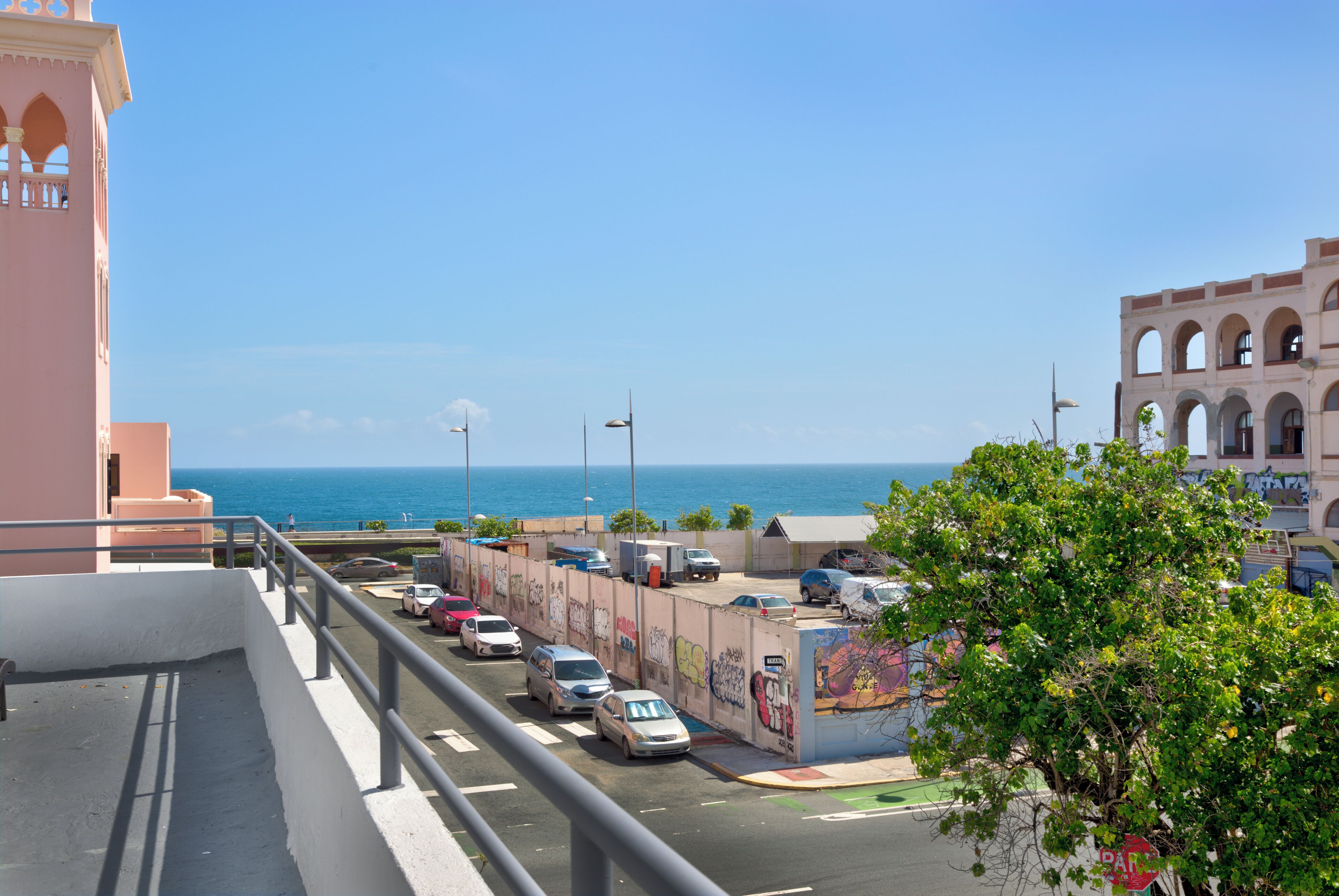 A coastal road with parked cars, a pedestrian walkway, and buildings with arched windows.