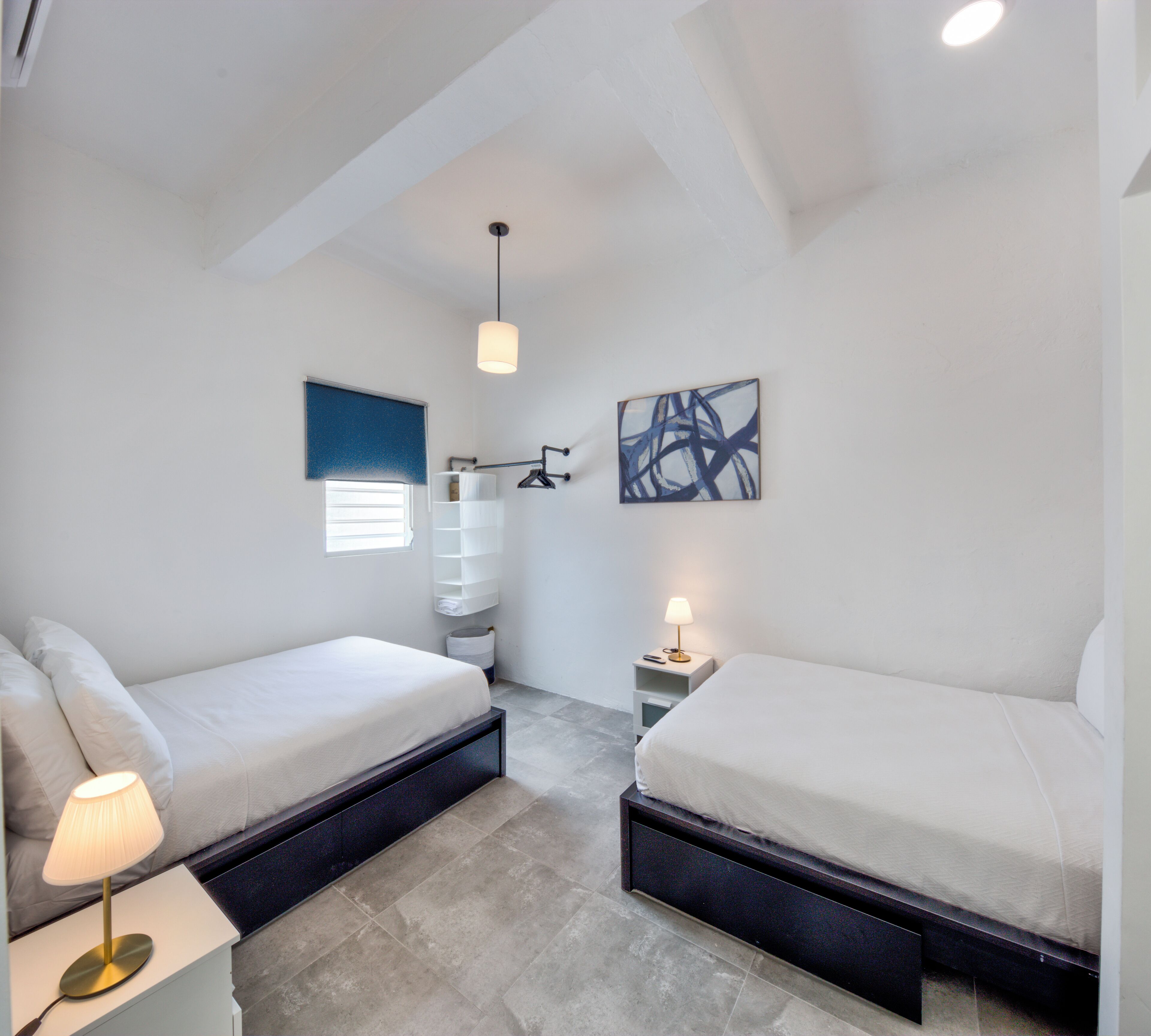 A modern bedroom with two beds, a bedside table with a lamp, a wall-mounted shelf, and a ceiling light.