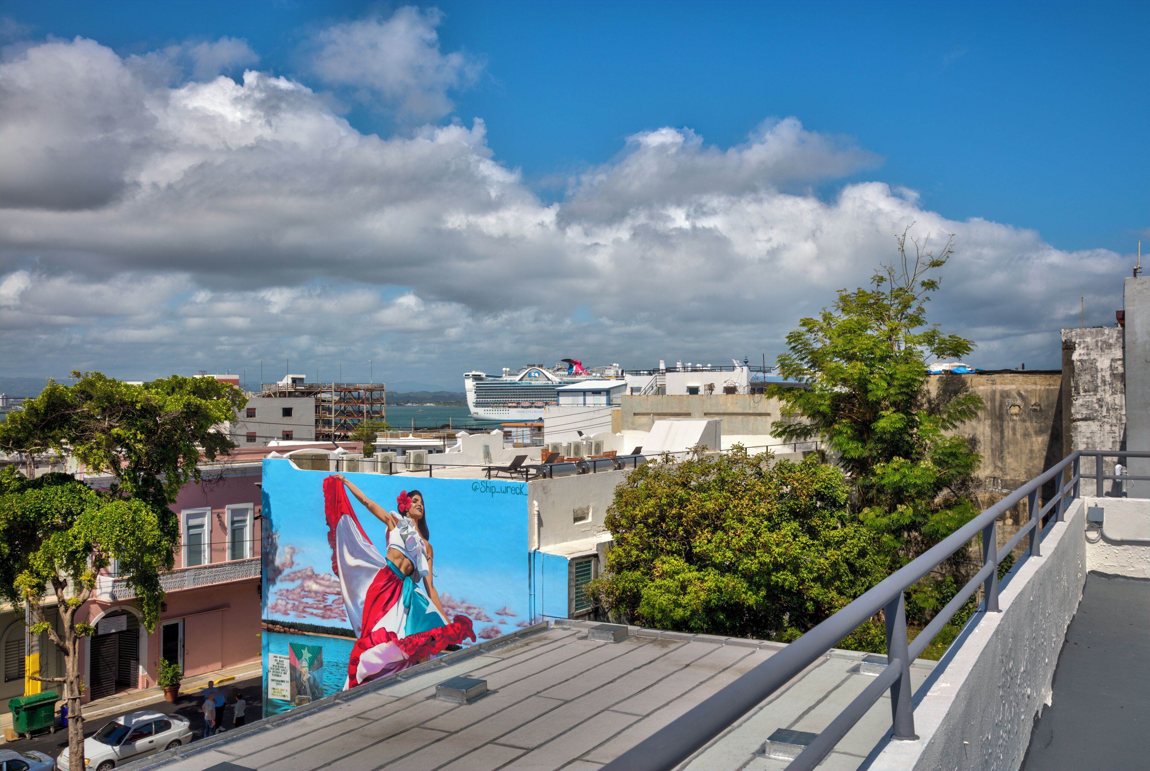 A cityscape with a large mural on a building, a cruise ship in the background, and a clear blue sky.