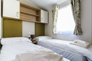 A hotel room with two beds, a wooden headboard, a bedside table with a lamp, and a window with curtains. - Caravan with stunning mountain views (Glencoe)