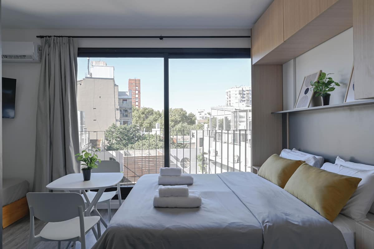 A modern hotel room with a large bed, a small table with chairs, a potted plant, and a view of the cityscape through large windows.