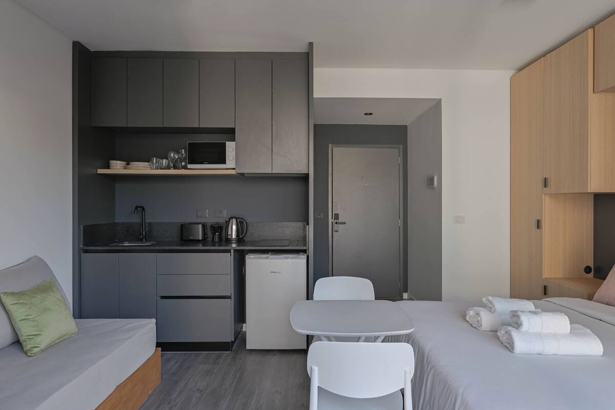A compact hotel room with a kitchenette, a small dining table, a bed with towels, and a sofa.
