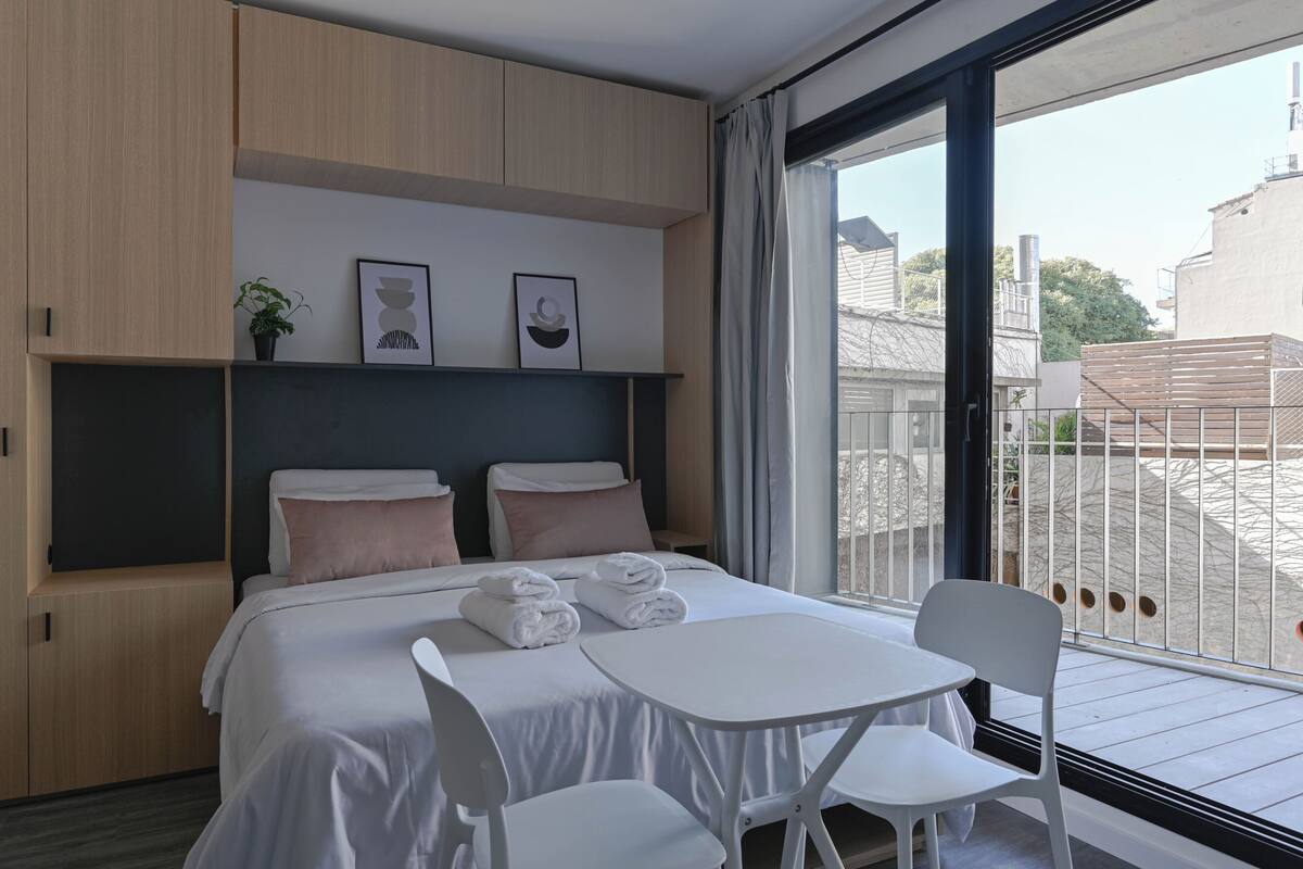 A modern hotel room with a bed, bedside tables, a small table with chairs, and a balcony with a railing.