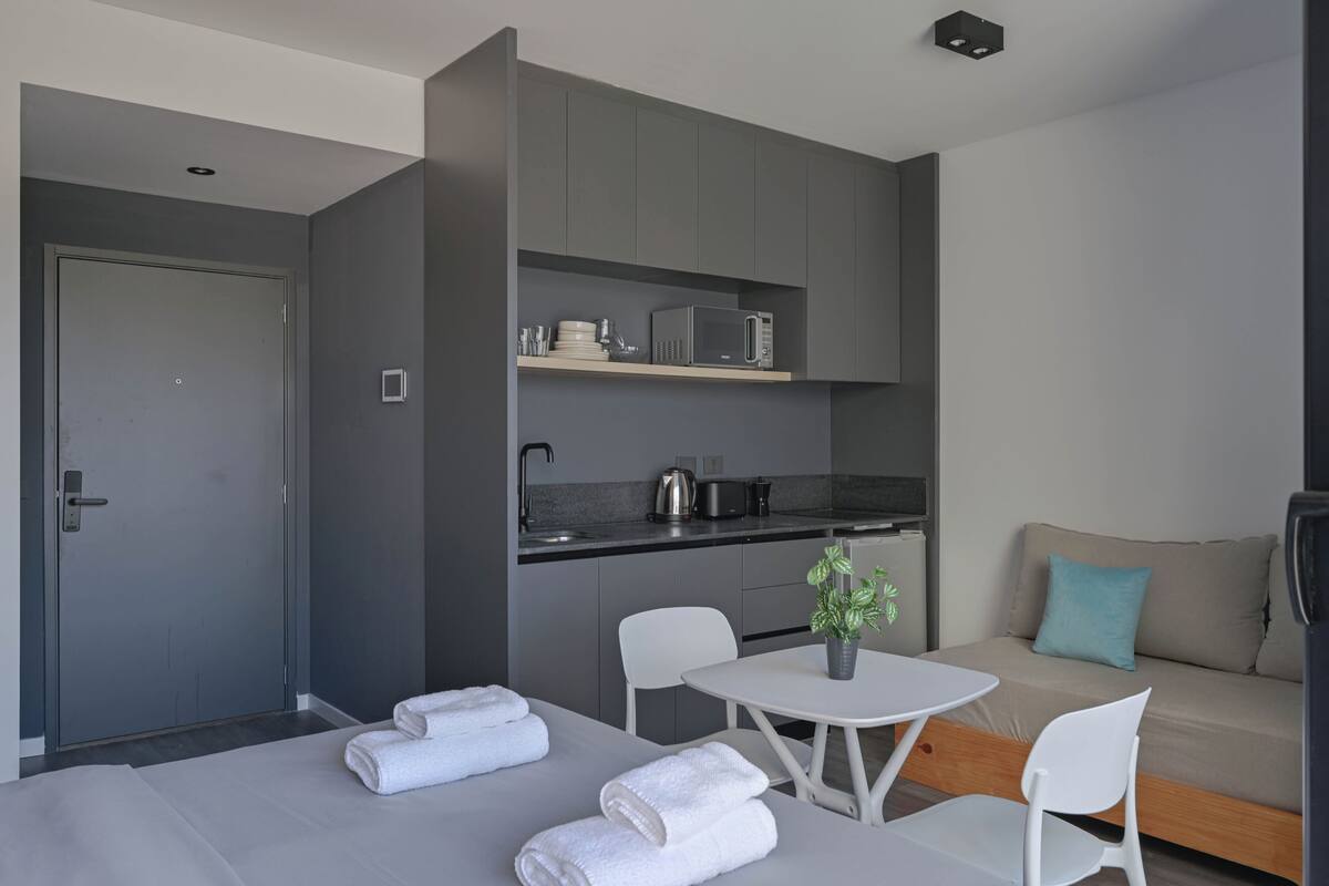 A modern hotel room with a kitchenette, a sofa, a dining table with chairs, and a bed with towels.