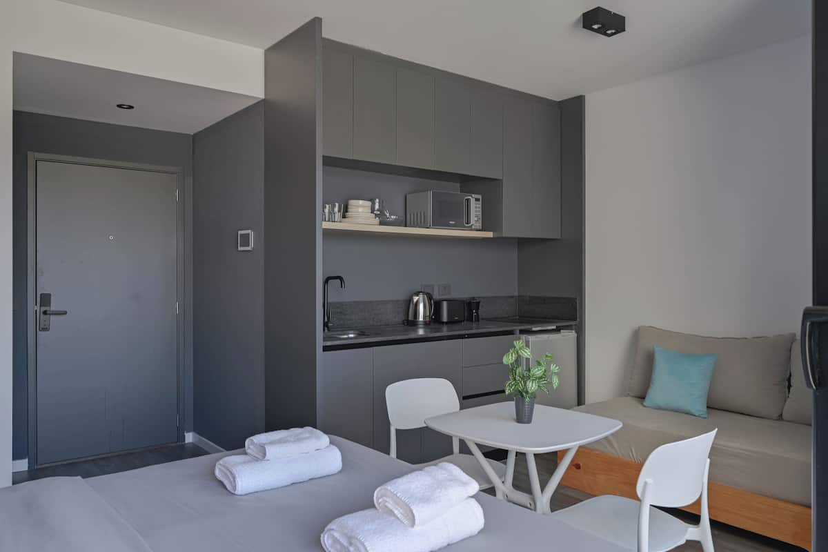 A modern hotel room with a kitchenette, a sofa, a dining table with chairs, and a bed with towels.