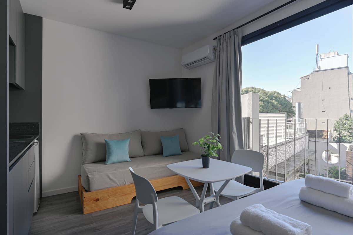 A modern hotel room with a sofa, a small table, a TV, and a balcony with a view of buildings.