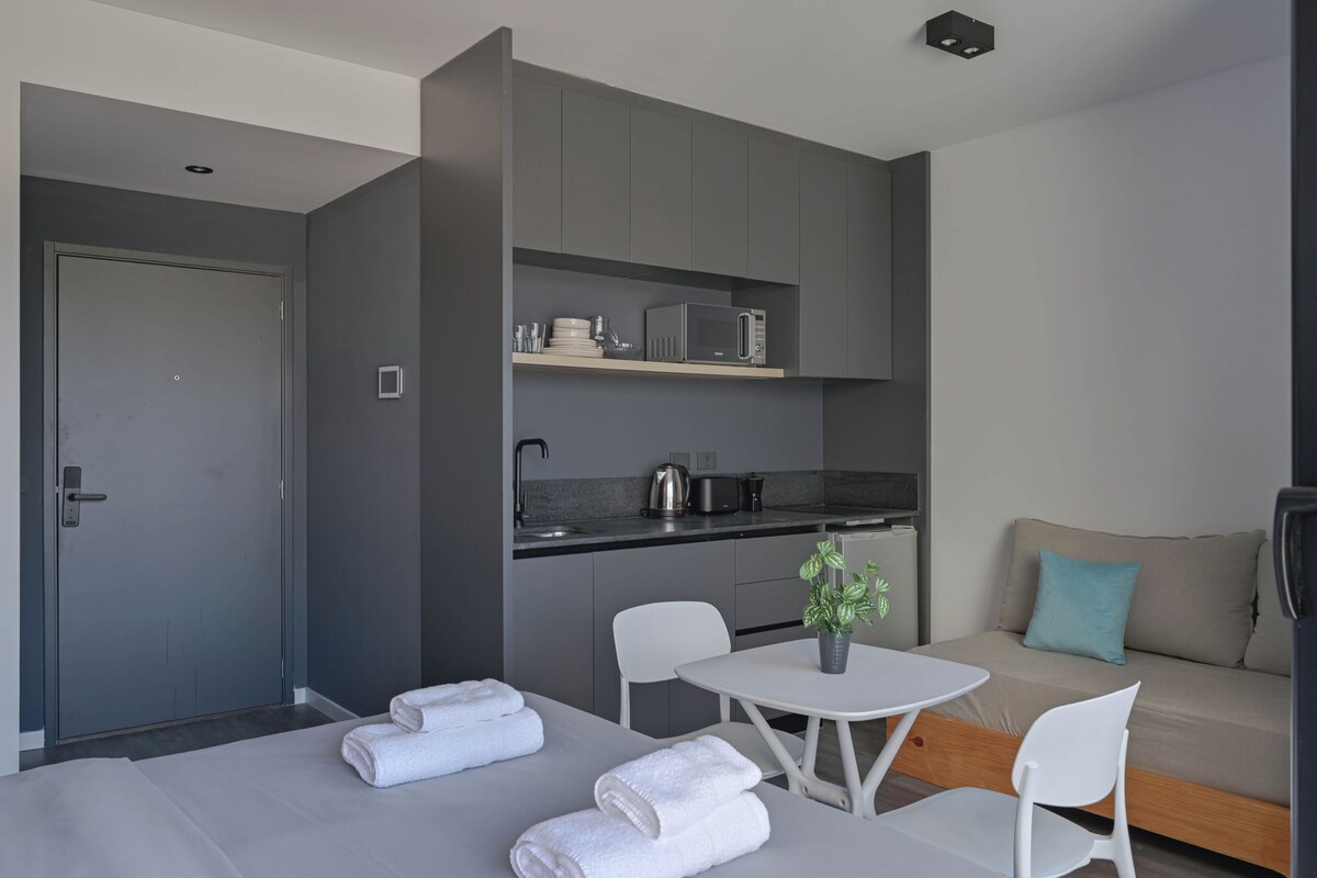 A modern hotel room with a kitchenette, a sofa, a dining table with chairs, and a bed with towels.