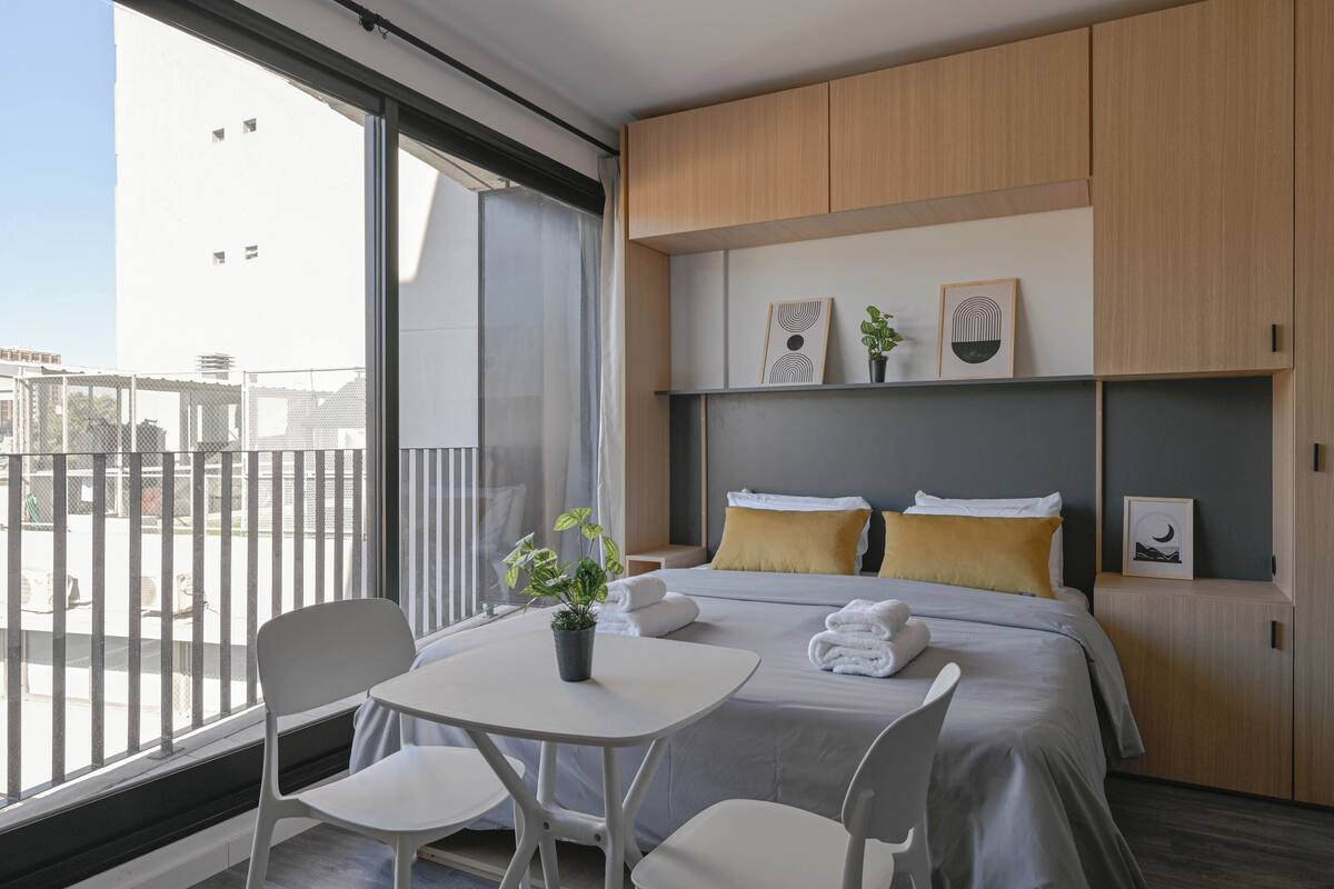 A modern hotel room with a balcony, a bed with two pillows, a small table with a plant, and a chair.