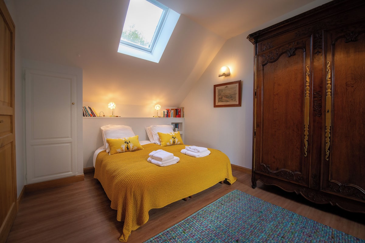 Ein Schlafzimmer mit Oberlicht, einem Bett mit gelber Bettwäsche, einem Holzschrank und einem kleinen Bücherregal.