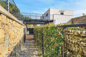 Ein schmaler Steinweg führt zu einem modernen Gebäude, darüber eine Metallbrücke.