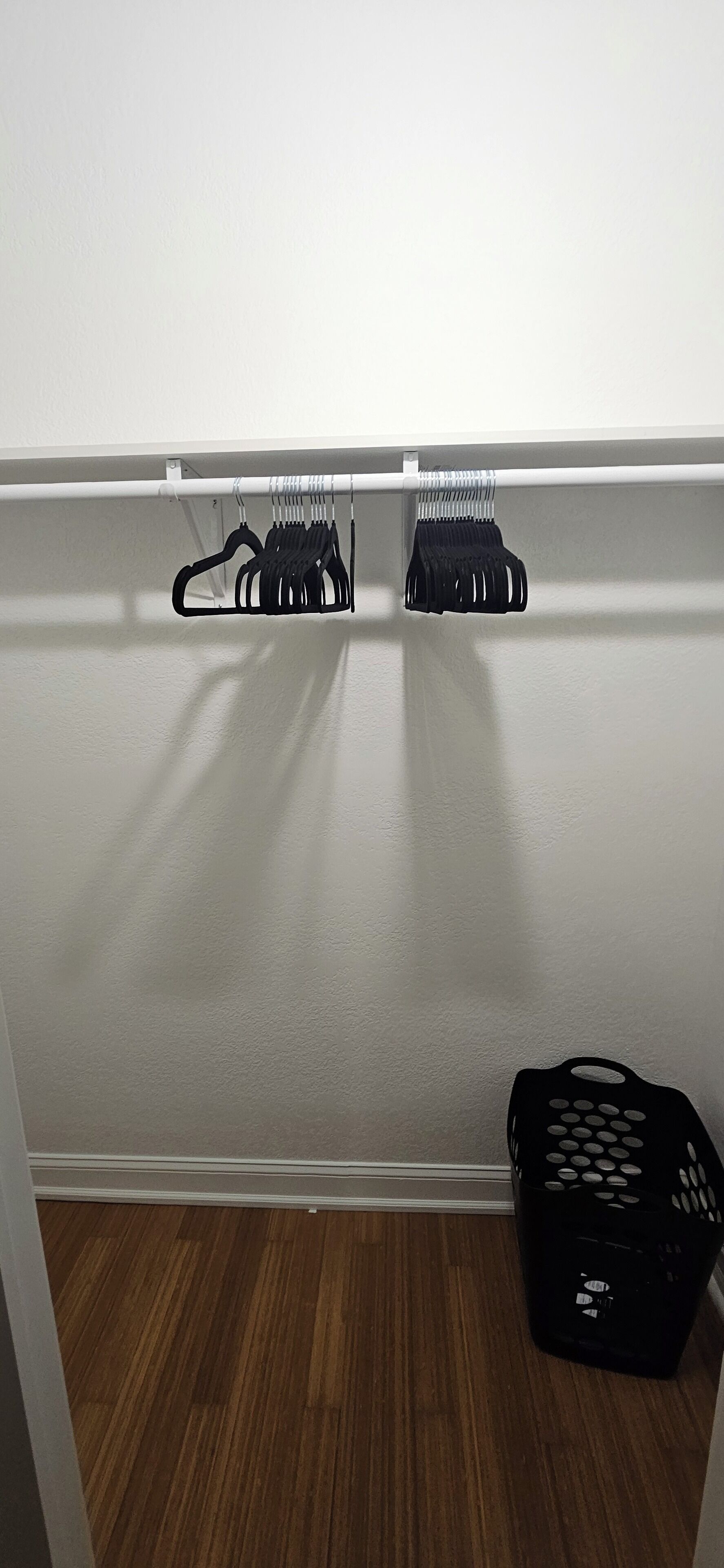 Vaatekaappi, jonka seinä on valkoinen ja lattia puuta. Vaatetangossa on kaksi mustaa henkaria. Lattialla on musta kori.