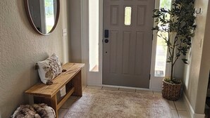 A hallway with a grey door, a bench with a cushion, a mirror, a rug, and a potted plant.