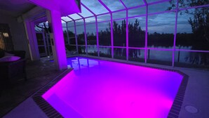 A glass-enclosed swimming pool with a view of a lake and trees, surrounded by a patio area with chairs.