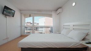 A hotel room with a bed, a TV mounted on the wall, and a balcony with a view of buildings and a Ferris wheel. - Charming Studio - Sleeps 2 - Close to the sea (Agde)