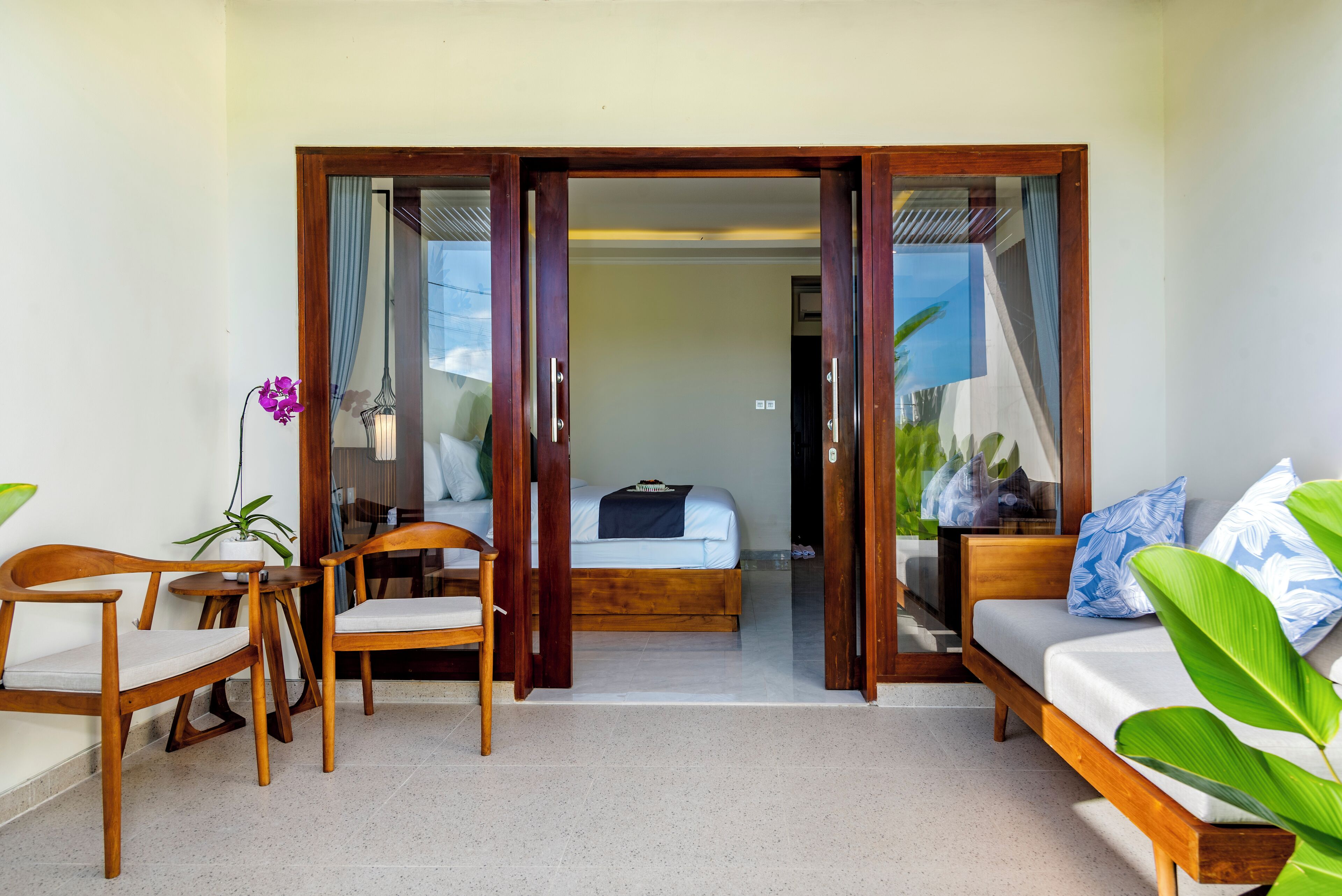 Habitación de hotel con cama, sofá, dos sillas de madera, una mesita y una planta en maceta.