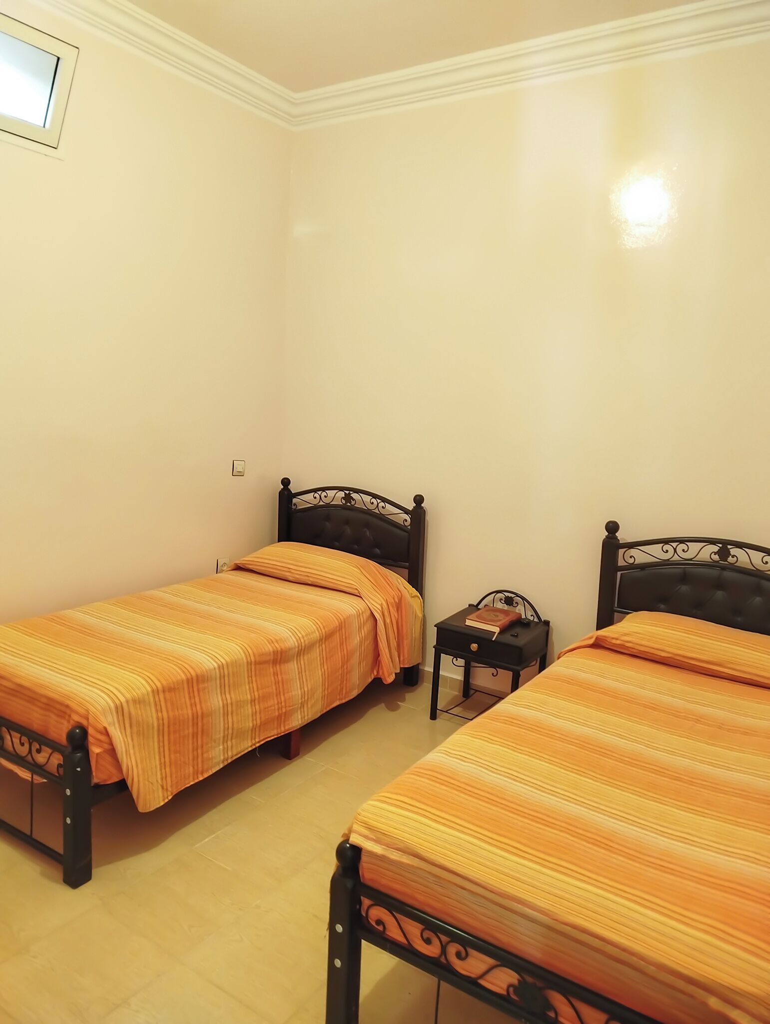 Een kamer met twee bedden, elk met een donker hoofdbord en beddengoed met oranje strepen. Er staat een klein nachtkastje met een boek erop.
